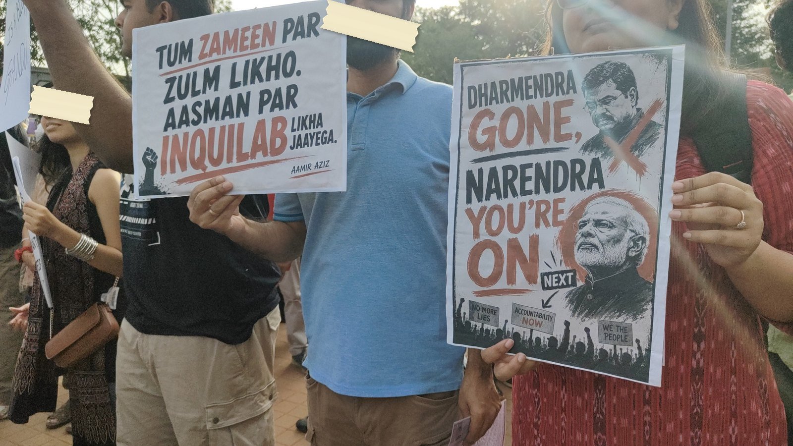 Two posters held side by side at Freedom Park, one quoting Aamir Aziz and one reading Dharmendra gone, Narendra you're on