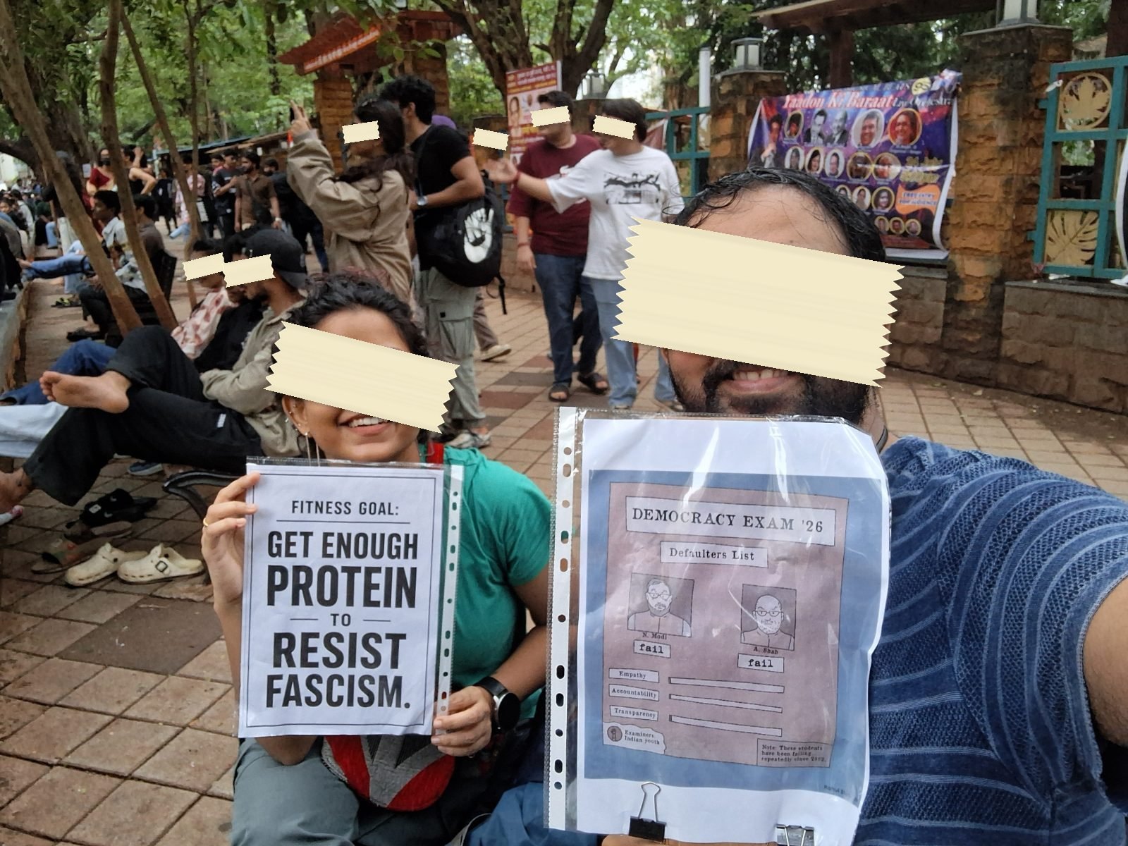 Two placards side by side: a fitness-goal joke about resisting fascism, and a mock answer sheet titled Democracy Exam 26 stamped fail