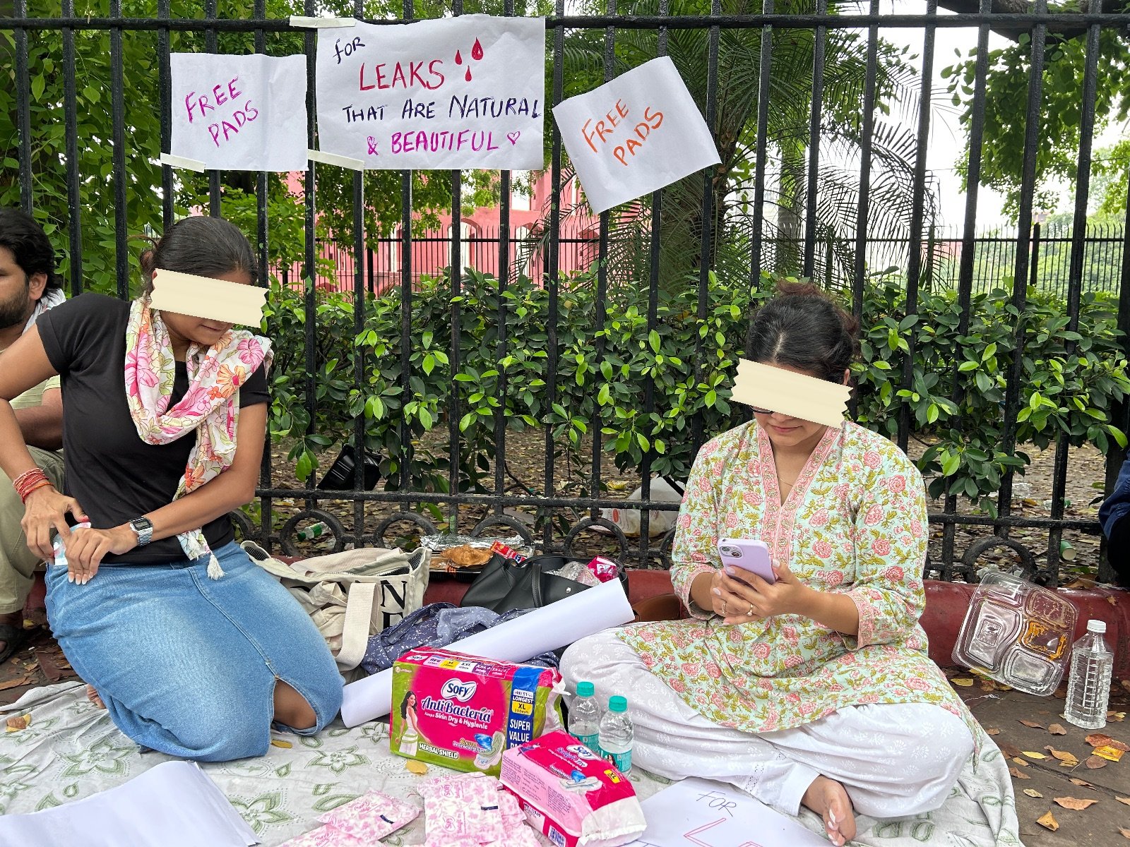 Two young women sitting on a mat at a protest camp with packets of sanitary pads laid out in front of them, and three hand-lettered signs tied to the railing behind reading FREE PADS, for LEAKS THAT ARE NATURAL AND BEAUTIFUL, FREE PADS
