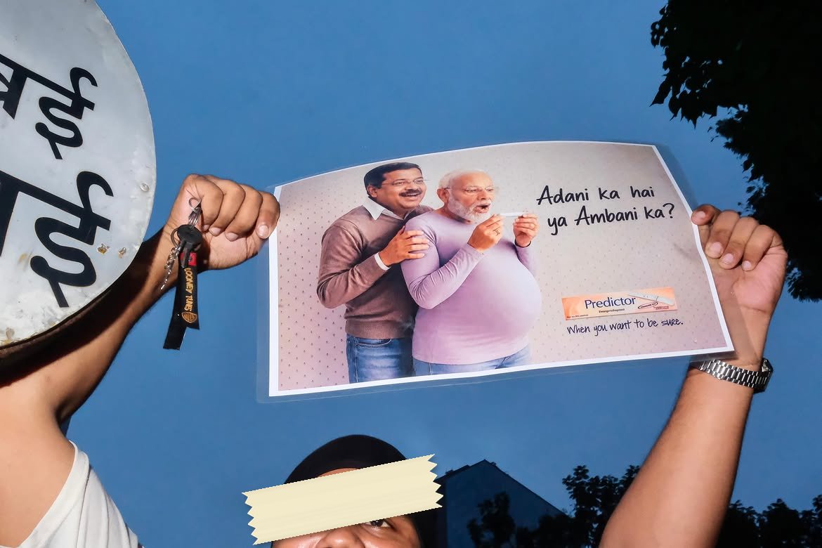 A landscape print held up at dusk by two hands, parodying a pregnancy-test advertisement. Two men stand together, one behind the other with his hands on his shoulders, the front one holding a test stick and looking down at it. The line reads Adani ka hai ya Ambani ka, and the Predictor logo sits at the bottom right above the words When you want to be sure.