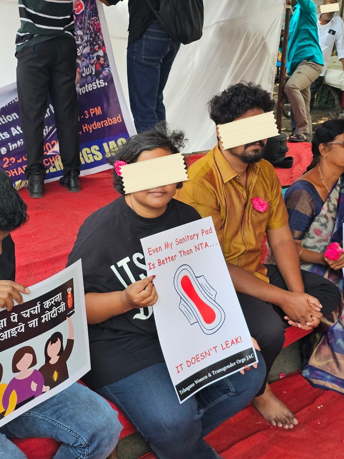 A sit-in on a red carpet in Hyderabad, a young woman holding a printed placard showing a drawn sanitary pad and reading Even my sanitary pad is better than NTA, it doesn't leak