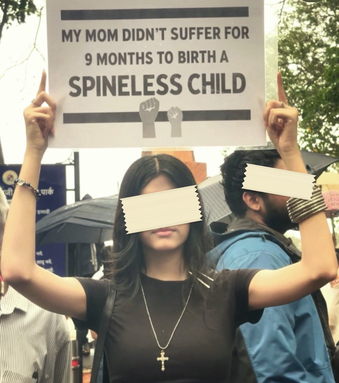 A printed placard held overhead in the rain reading MY MOM DIDN'T SUFFER FOR 9 MONTHS TO BIRTH A SPINELESS CHILD, with two raised-fist icons beneath the words