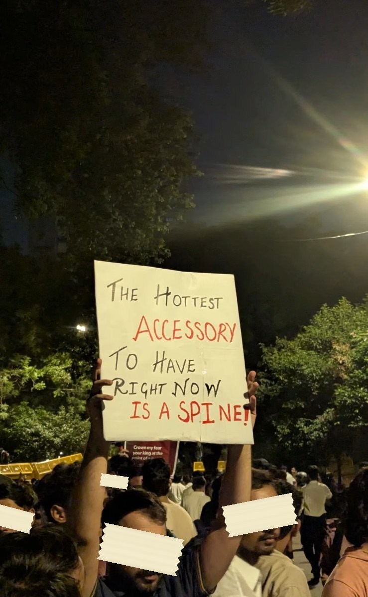 A hand-lettered placard held up at night reading THE HOTTEST ACCESSORY TO HAVE RIGHT NOW IS A SPINE, with ACCESSORY and SPINE written in red