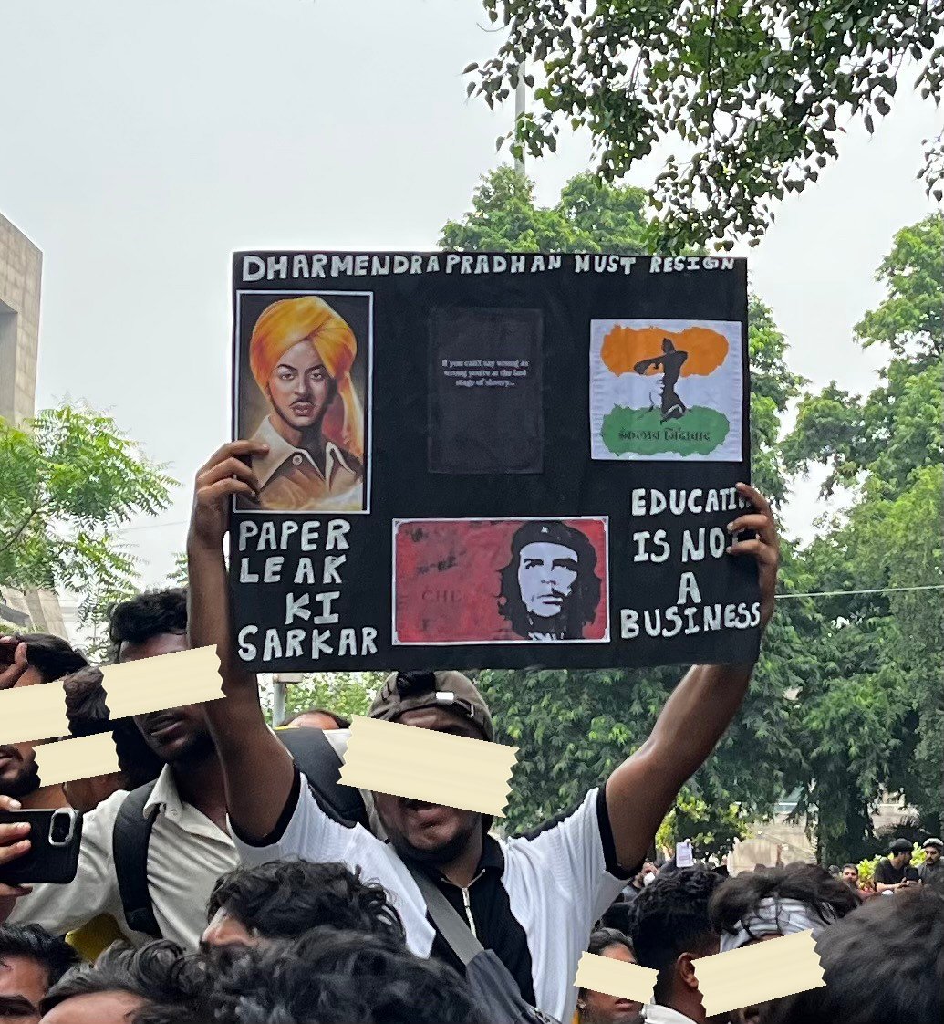 A hand-painted black board carrying portraits of Bhagat Singh and Che Guevara, reading Dharmendra Pradhan Must Resign, Paper Leak Ki Sarkar, Education Is Not A Business
