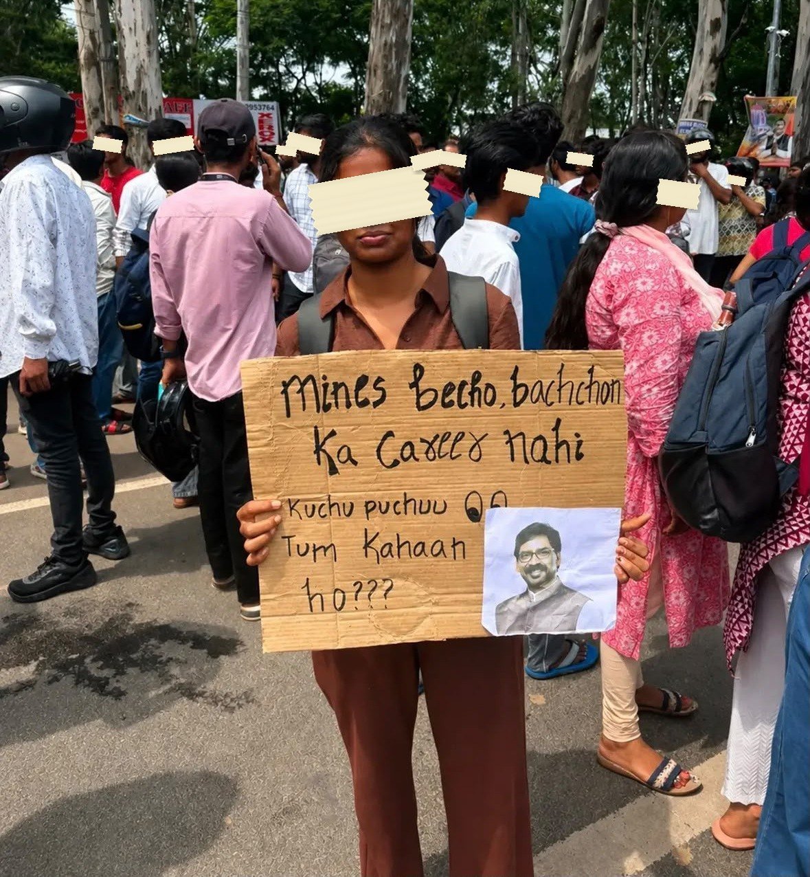 A young woman holds a brown cardboard placard reading Mines becho bachchon ka career nahi, with a cut-out printed portrait of a man pasted into the lower right corner.