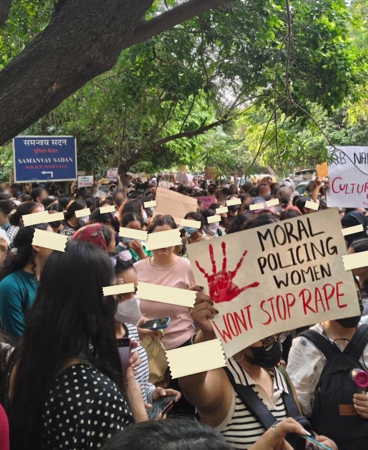 A hand-lettered placard held above a dense crowd on a Delhi street, a red painted handprint beside the words MORAL POLICING WOMEN WONT STOP RAPE.