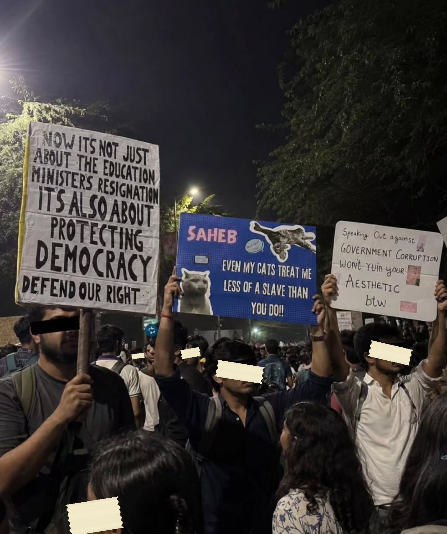 Three placards held up at night in a crowd. On the left, hand-lettered in black capitals on white: NOW ITS NOT JUST ABOUT THE EDUCATION MINISTERS RESIGNATION ITS ALSO ABOUT PROTECTING DEMOCRACY DEFEND OUR RIGHT. In the middle, a printed blue board addressed to SAHEB carrying two photographs of cats and the line EVEN MY CATS TREAT ME LESS OF A SLAVE THAN YOU DO. On the right, hand-lettered and decorated with small stickers: Speaking Out against GOVERNMENT CORRUPTION won't ruin your Aesthetic btw.