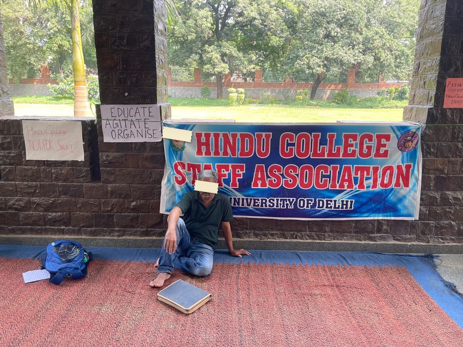 A man sitting cross-legged on a dhurrie in a college portico, in front of a printed vinyl banner reading HINDU COLLEGE STAFF ASSOCIATION, University of Delhi, with his shoes to one side and a hardbound book face-down on the mat beside him.