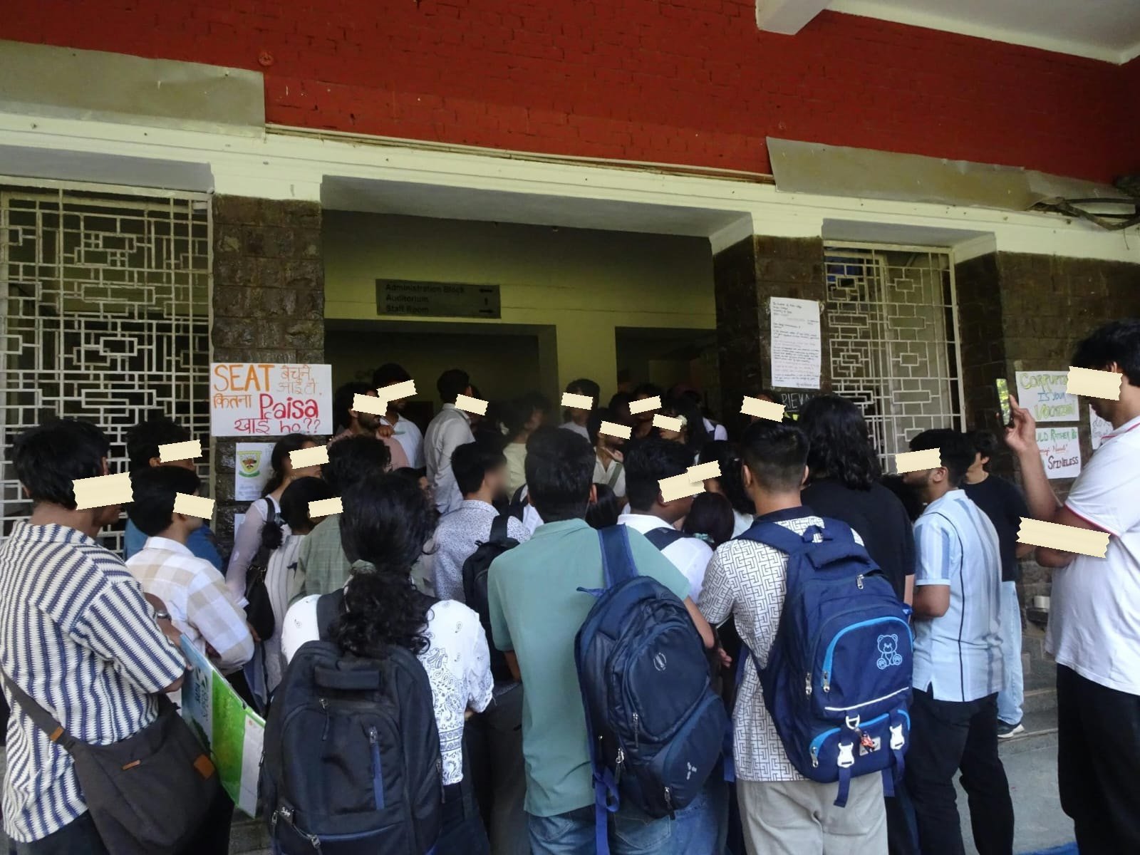 A crowd of students pressed around the doorway of the administration block, with a hand-lettered sign beside the door reading SEAT बेचने आई तो, कितना Paisa खाई हो?? and another reading CORRUPTION IS YOUR VOCATION.