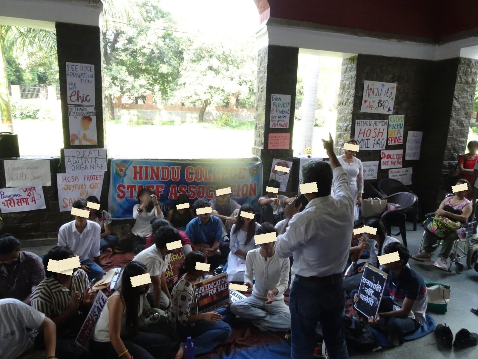 A sit-in under the arches of a college portico, students and staff seated on mats in front of the Staff Association banner, with hand-lettered placards taped to the pillars around them.