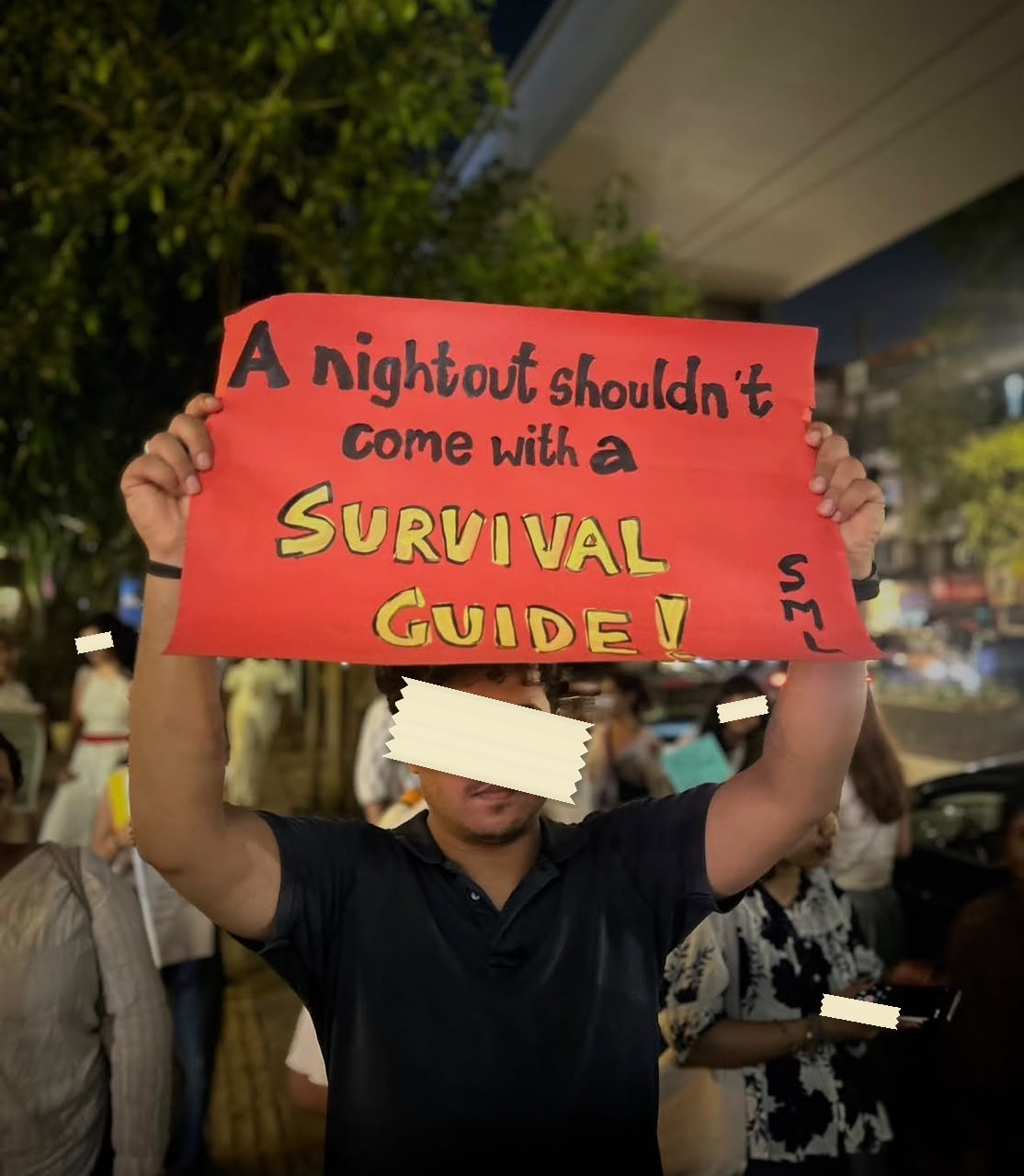 A red placard held overhead at night reading A nightout shouldn't come with a SURVIVAL GUIDE! with the letters SML in the corner.