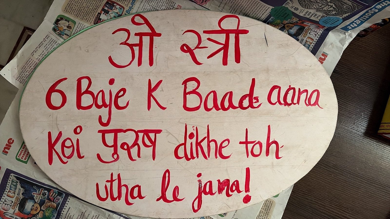 An oval board painted in red on white, photographed indoors on newspaper, reading in Hindi and Roman script: O Stree, 6 Baje K Baad aana, Koi purush dikhe toh utha le jana!