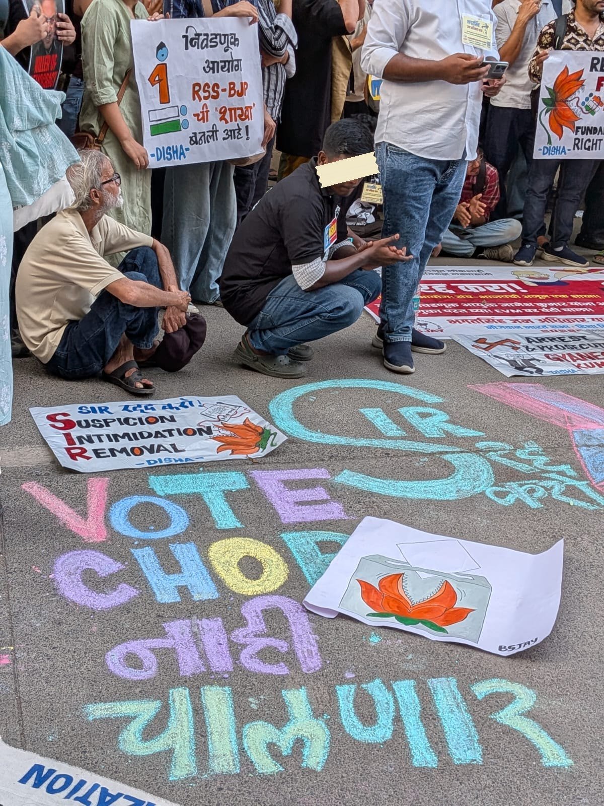 Chalk writing on tarmac reading VOTE CHORI nahi chalnar in Marathi, with the letters SIR drawn large in cyan outline, and printed placards lying on the road beside it.