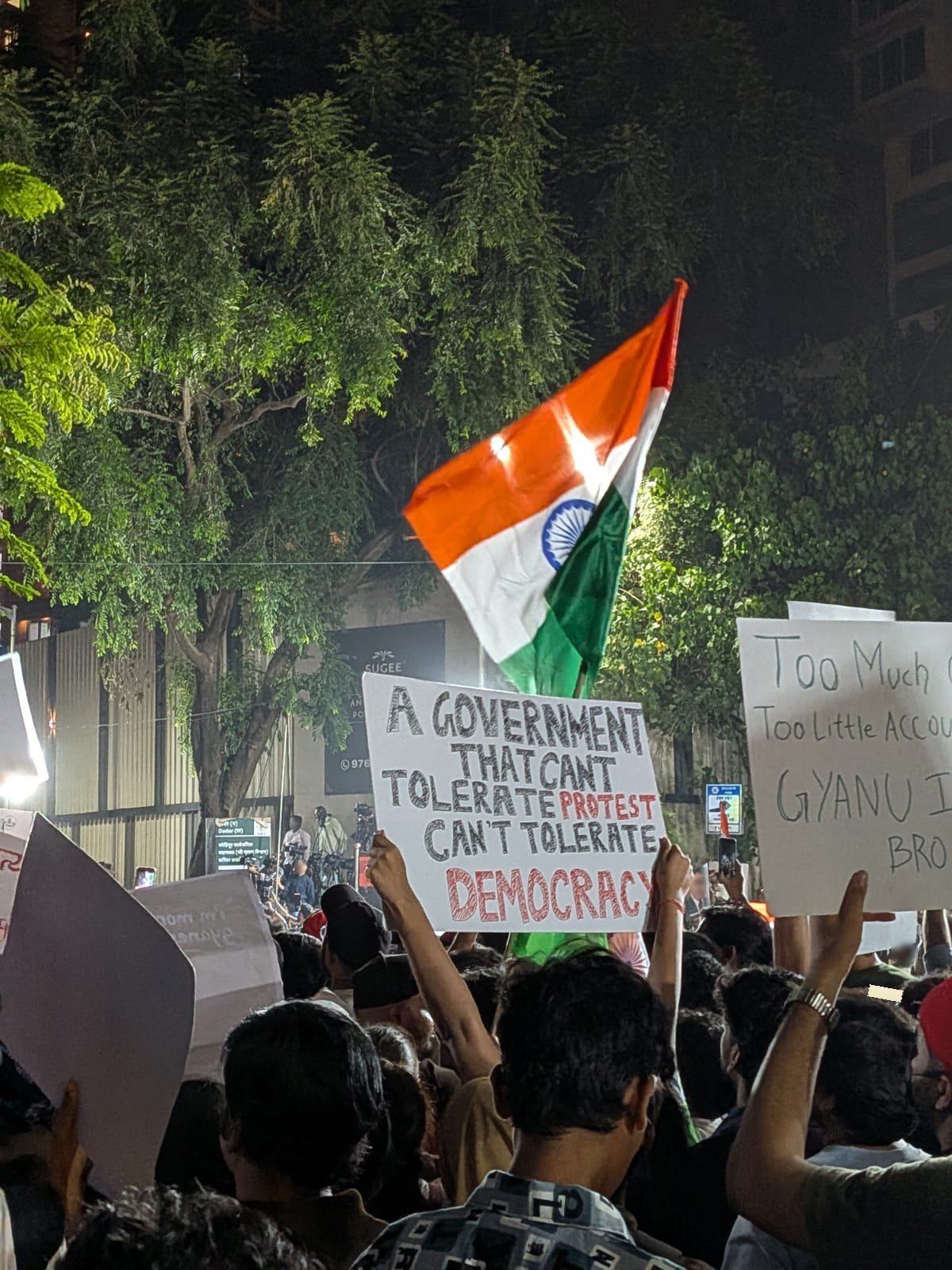 A night crowd with a large tricolour and a hand-lettered placard reading A GOVERNMENT THAT CAN'T TOLERATE PROTEST CAN'T TOLERATE DEMOCRACY.