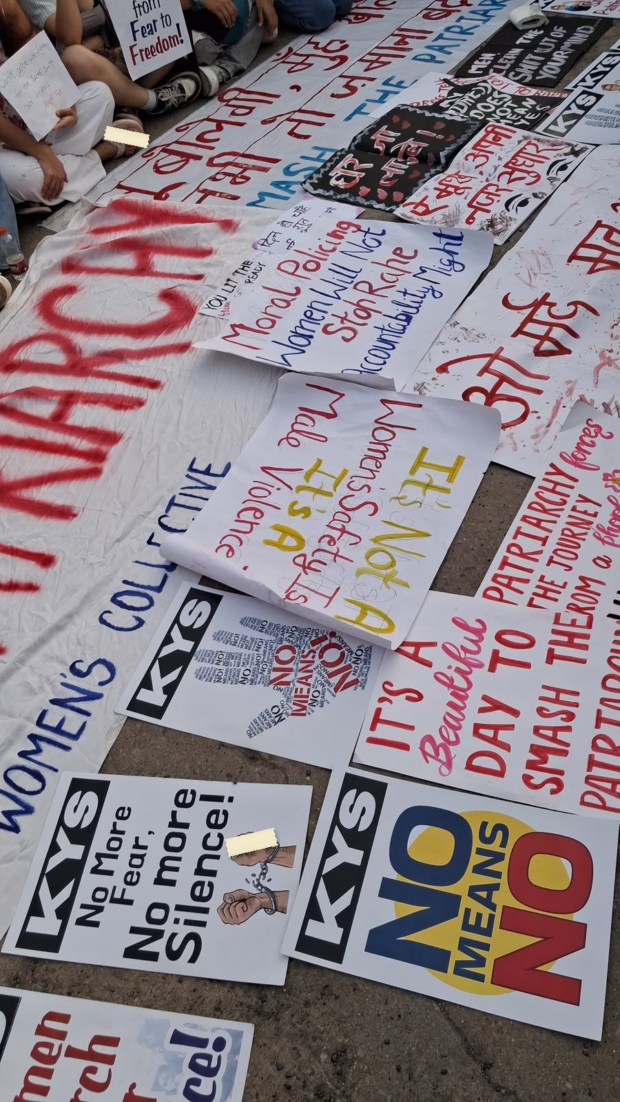 Dozens of placards laid flat on tarmac before a march: printed ones reading NO MEANS NO and No More Fear, No more Silence among hand-lettered sheets in several colours.