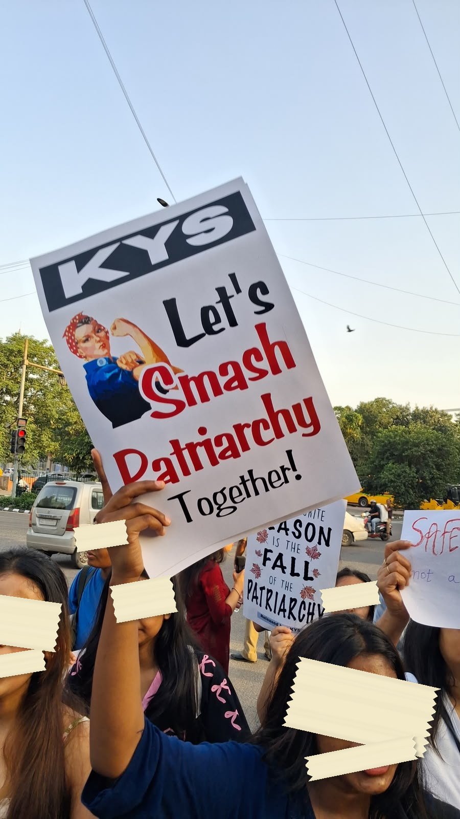 A printed placard held above a crowd: a black KYS masthead, a drawing of a woman flexing her arm, and the words Let's Smash Patriarchy Together.