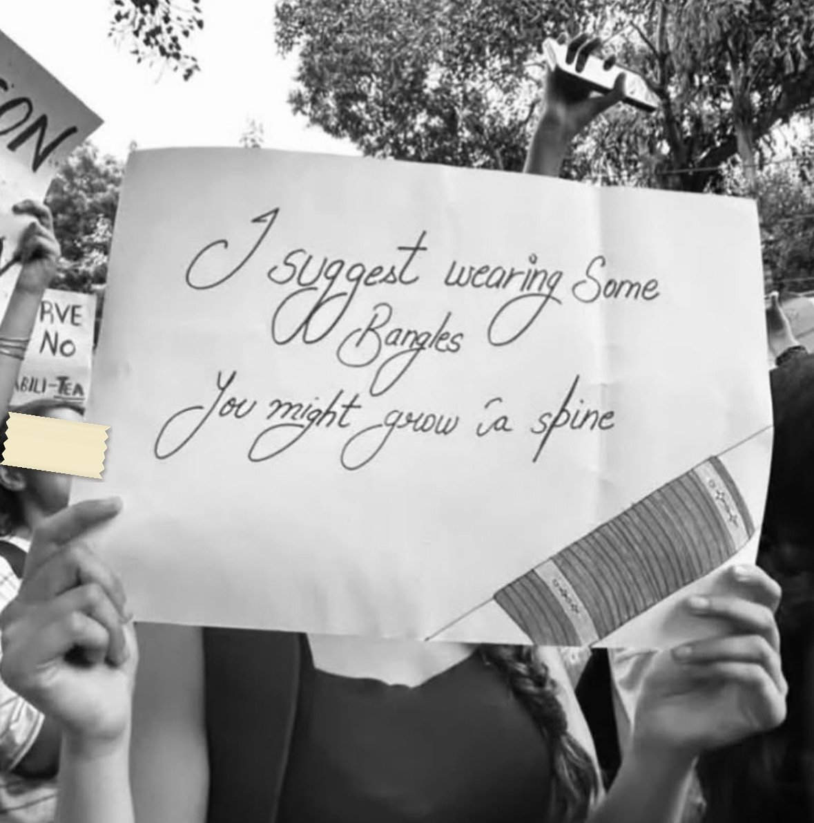 A hand-lettered placard in cursive: I suggest wearing some Bangles, You might grow a spine, held up to cover the face of the person carrying it.