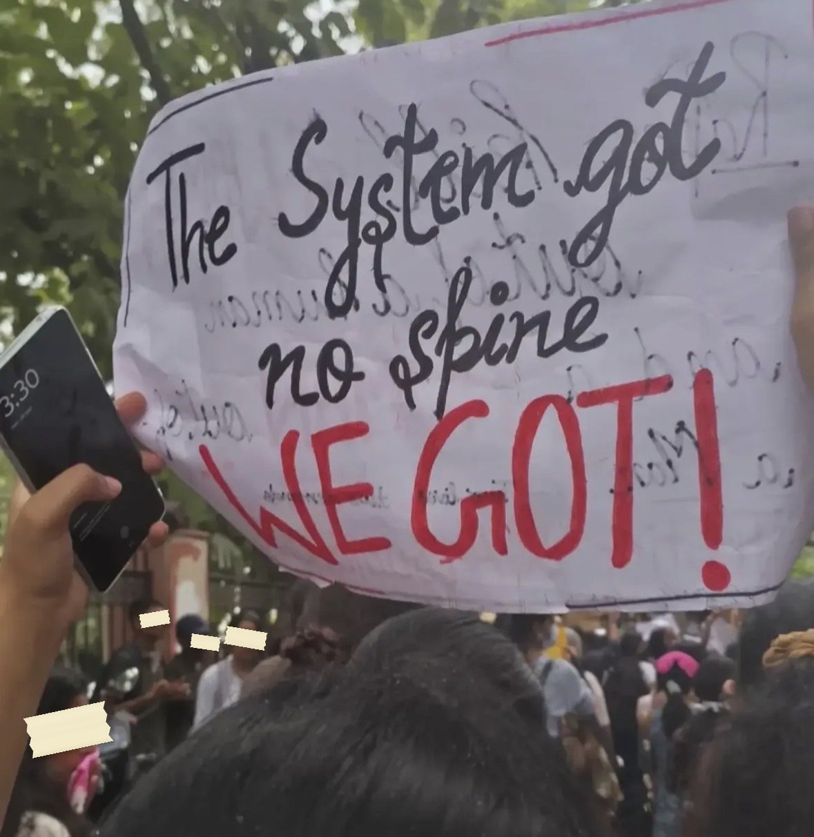 A placard reading The System got no spine in black, with WE GOT written beneath it in red capitals and an exclamation mark.