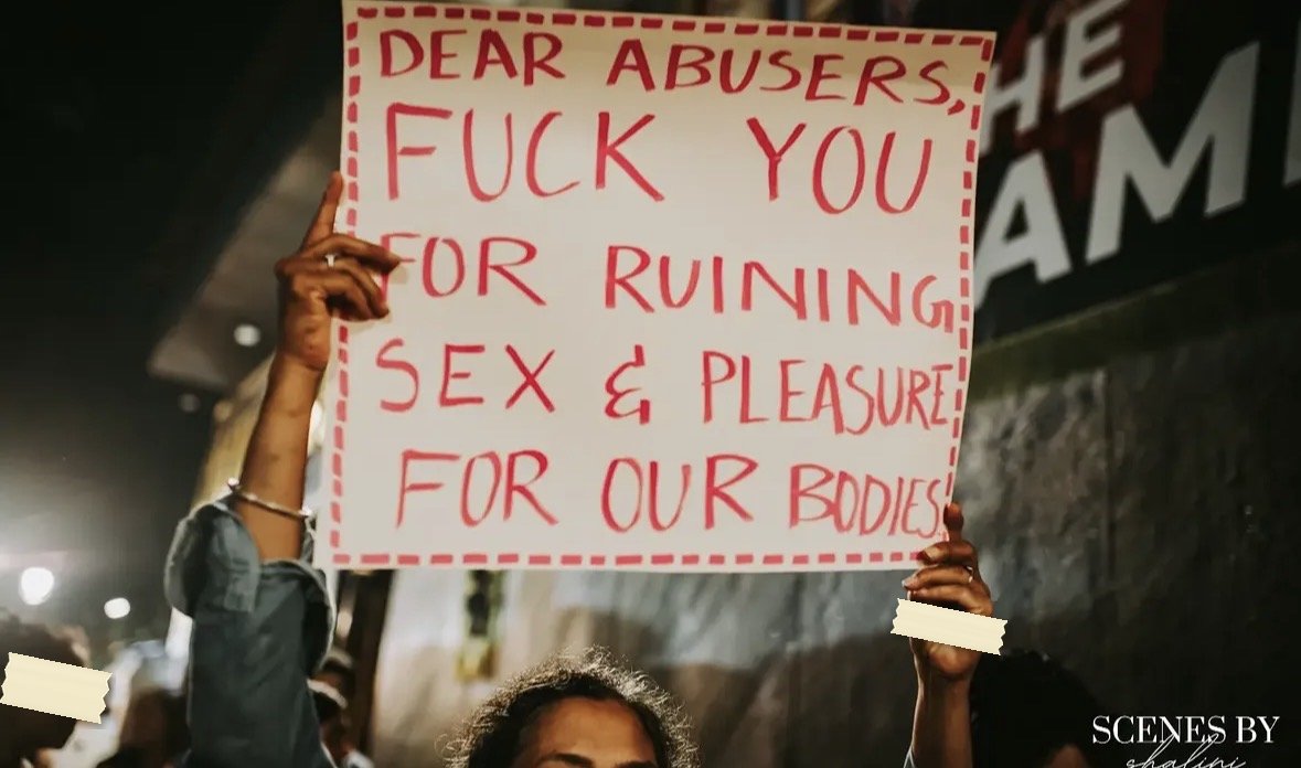 A pink-bordered placard at night reading DEAR ABUSERS, FUCK YOU FOR RUINING SEX AND PLEASURE FOR OUR BODIES.