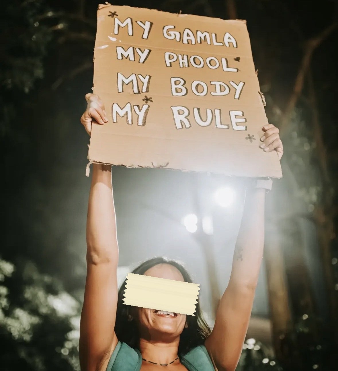 A cardboard placard held overhead at night, lit from behind, reading MY GAMLA MY PHOOL MY BODY MY RULE in white block capitals.