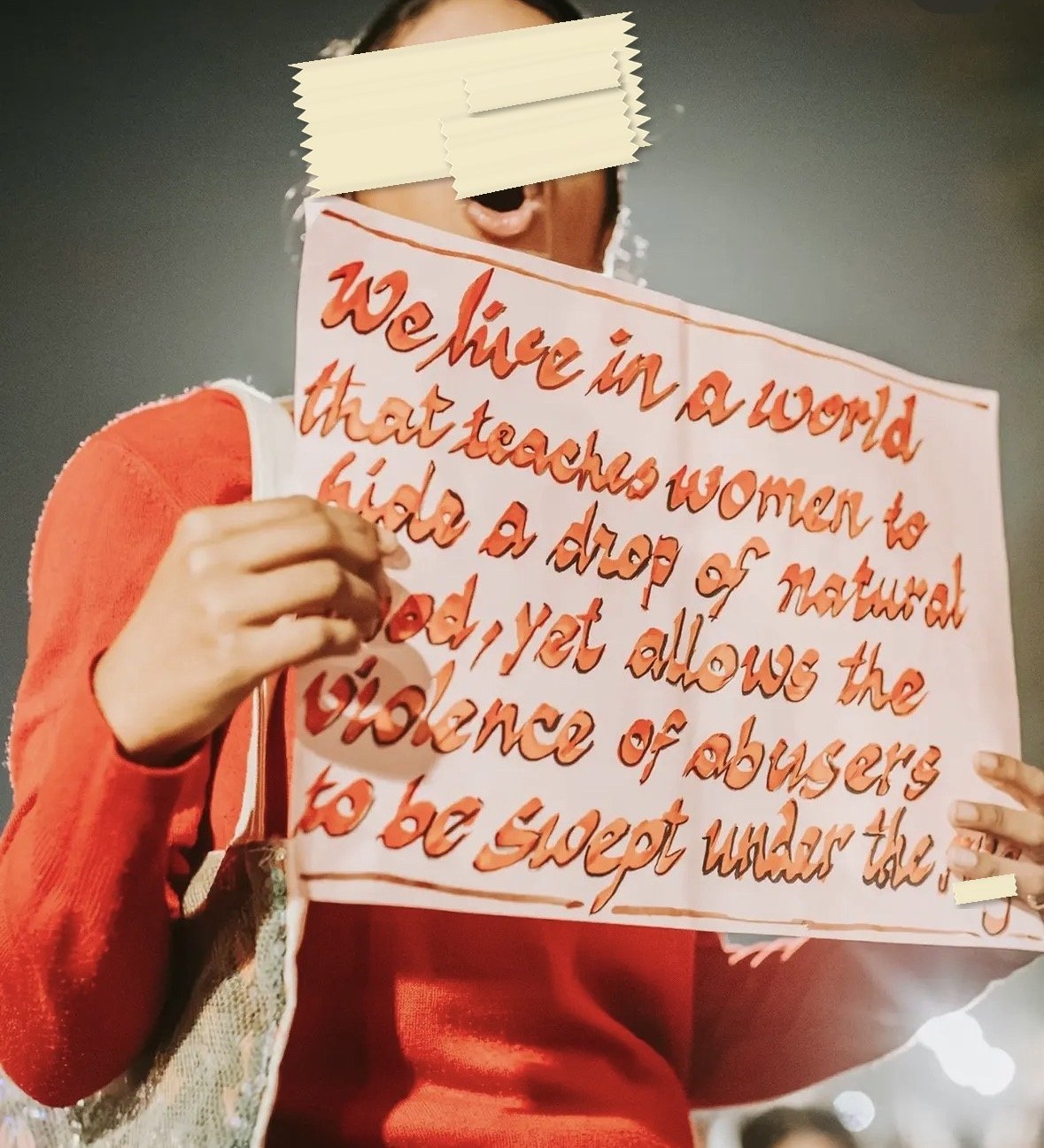 A placard in orange cursive: We live in a world that teaches women to hide a drop of natural blood, yet allows the violence of abusers to be swept under the rug.