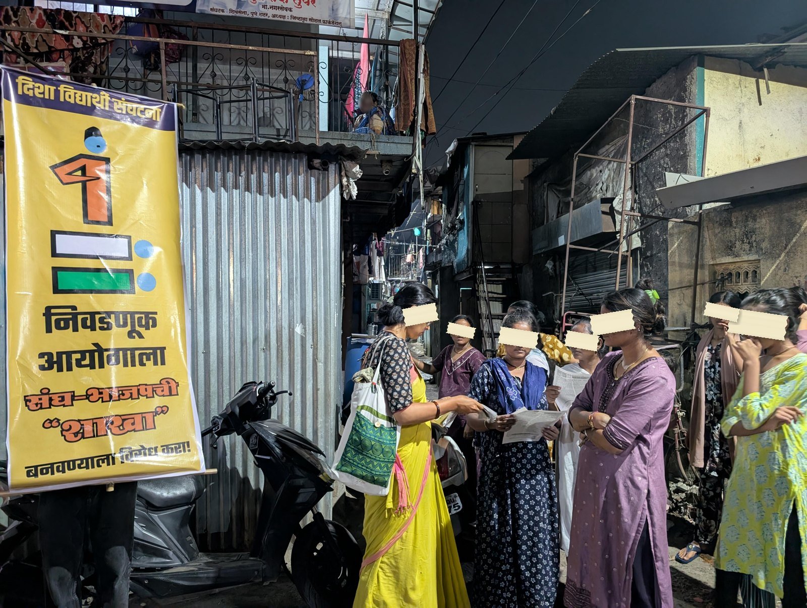 A night lane in Pune. A yellow printed banner with a ballot box stands against a corrugated shutter while a group of women read a leaflet together under a streetlight.