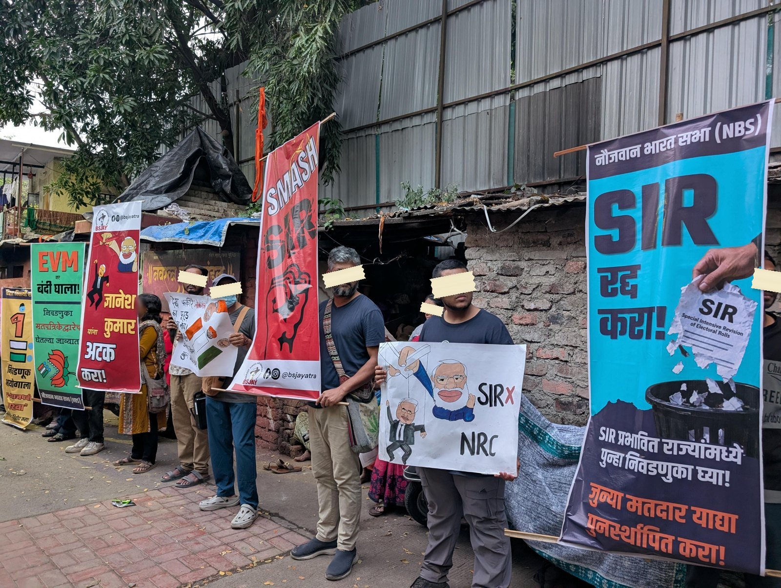 A row of banners propped along a corrugated wall in daylight, including a printed SIR radd kara banner, a red SMASH banner, and a hand-drawn sheet reading SIR crossed out above NRC.