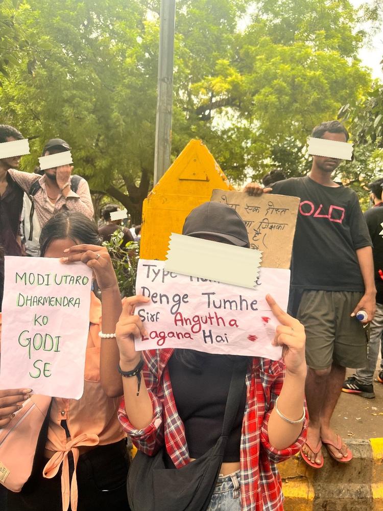 Two hand-lettered placards by a yellow barricade, one reading Modi utaro Dharmendra ko godi se, the other Type hum kar denge tumhe sirf angutha lagana hai.